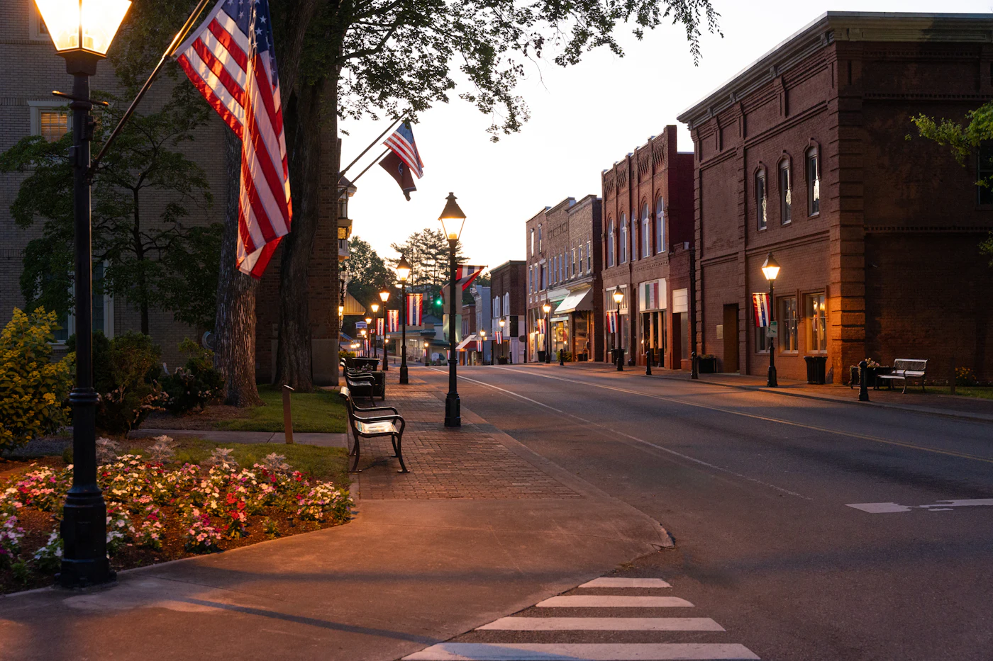 A quiet small-town main street at dusk, with brick storefronts, lamp posts and American flags